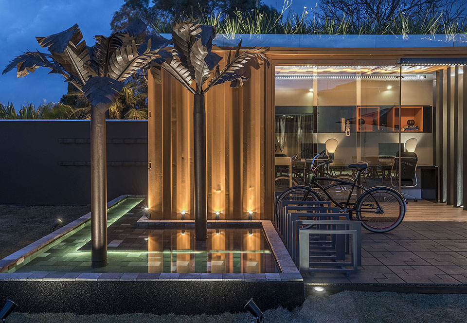 Bruna Bonfante e Juliana Lima ousaram na hora de projetar o Contêiner Office da CASACOR Minas Gerais 2015. Sustentável e elegante, o espaço apostou em recursos como: teto verde, piso reciclado, bicicletário e deck com espelho d’água. O fechamento em vidro garantia a continuidade entre o interior e o exterior da edificação. A construção foi pensada para ser itinerante, atendendo a necessidade de empresas que precisam de espaços temporários em cidades distantes de sua sede. Feita a partir de um contêiner de 40 pés, a estrutura reúne uma sala de reunião, uma sala do diretor e uma copa.