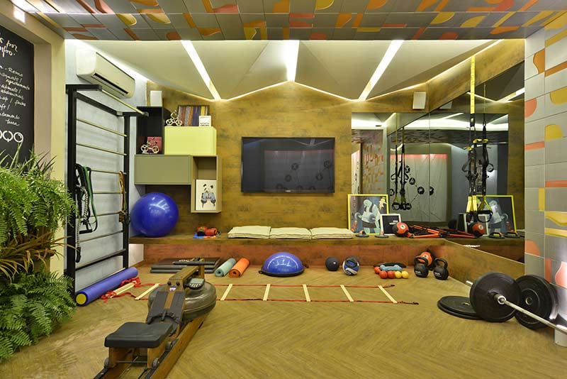 Fitness da Casa - Rita Braga . O painel da TV é inteiramente revestido com MDF aço corten, de aparência oxidada, que também serve de base para espelhos em L que permitem observar a execução dos exercícios. Nichos e basculantes que combinam tons neutros e de amarelo possuem profundidades e dimensões distintas, em um exercício de volumetria. Um banco também ocupa toda a extensão da parede, que auxilia no apoio de materiais e também é feito para descansar, sobre futons de tecidos crus. Destaque ainda para o piso aplicado no formato espinha de peixe, servindo de inspiração para a construção do teto - que deixa a luz passar entre as frestas.