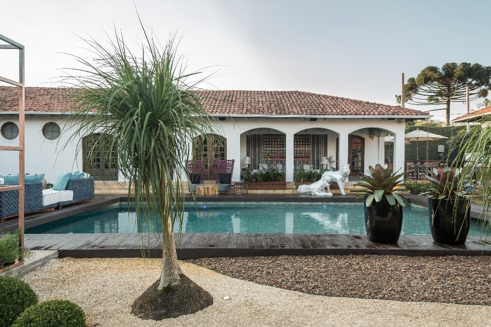 CASACOR Paraná 2018. Jardim e Piscina - Guto Ciccarino e André Derkcz. Como em um retrato a céu aberto, o olhar percorre os 386m² e desvenda aos poucos as plantas exóticas e a piscina com borda de pedras vulcânicas. No mobiliário, destaque para o uso do tricô com fios sintéticos, resistente ao clima.