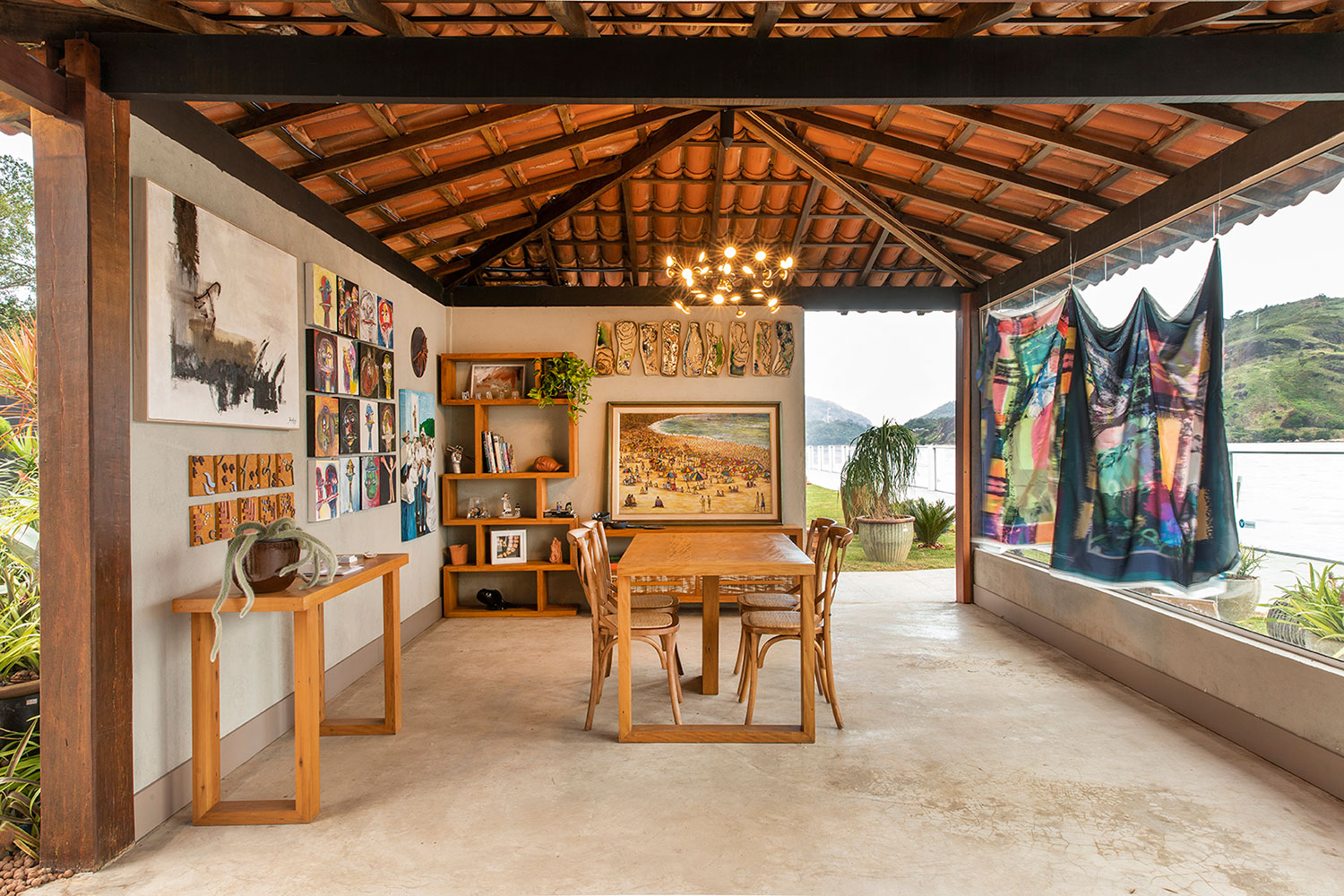 Casa Viva - Carol Zanetti Campanharo. O ambiente de 90 m² é inspirado em um ateliê e dedicado ao Sebrae-ES. Telas, gravuras e instalações de 12 artistas capixabas interagem com a natureza, emoldurada pela fachada em vidro. O espaço contemporâneo, com chão e paredes neutralizados com cimento queimado, também reverencia o estilo colonial no telhado reaproveitado do Clube Álvares Cabral.