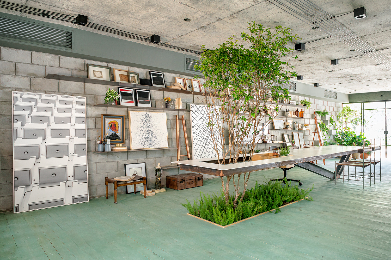 CASACOR Bahia. Oficina do Artista - GAM Arquitetos. No espaço de 110 m2, o concreto aparente da laje e dos pilares originais - antes coberto por gesso e tinta - recupera seu protagonismo. Guido Ramos, Anna Fernandes e Marcelo Bezerril também desenharam a mesa em aço com quase 6 m de comprimento, um único apoio no piso e extensão em balanço. Ela abraça a jabuticabeira plantada no local. No mobiliário, um mix de peças de design nacional, de Gustavo Bittencourt, Sérgio Rodrigues e Jader Almeida.