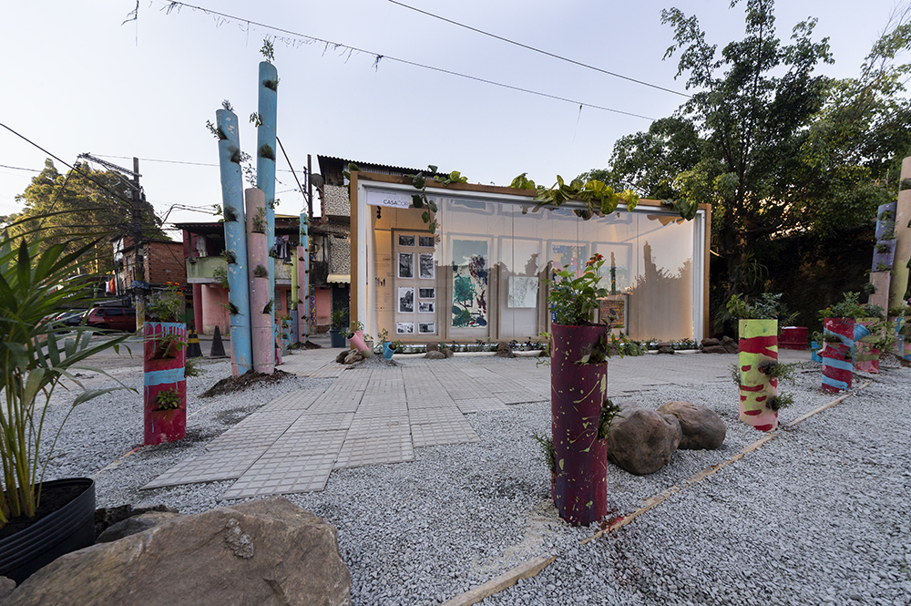 Vista externa da Galeria. Totens coloridos com plantas. Contêiner com obras de arte expostas no centro