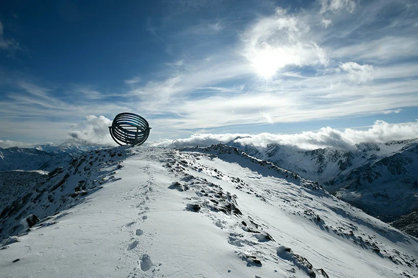 Montanha com neve e céu azul. No topo, estrutura artística