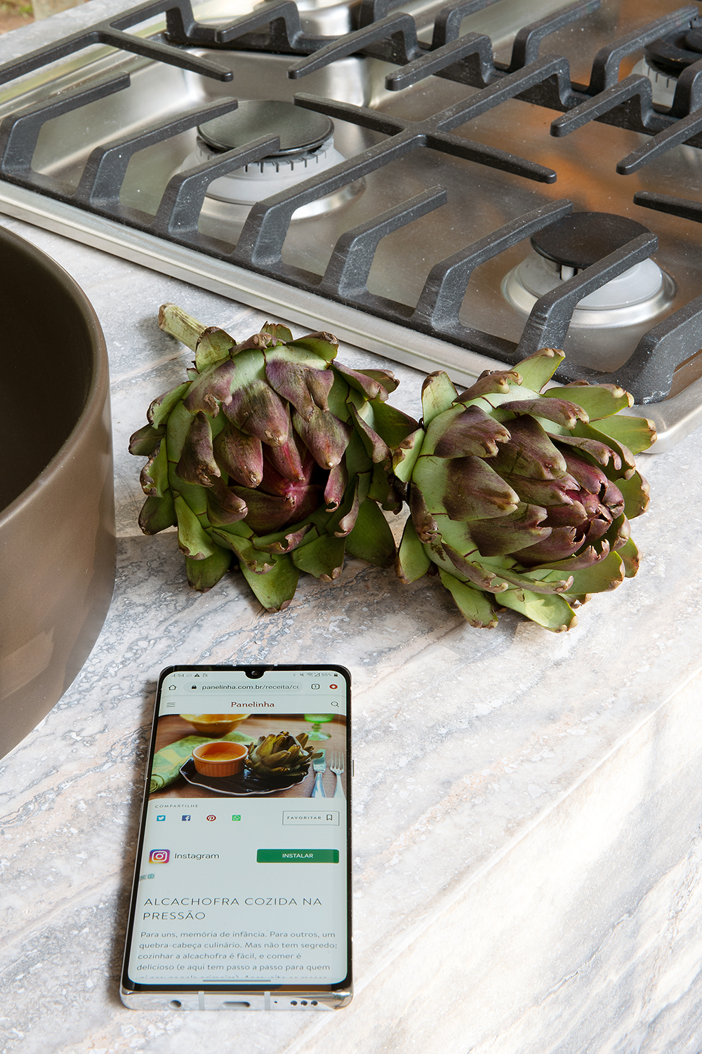 Bancada da cozinha com celular e alcachofras em primeiro plano e cooktop ao fundo