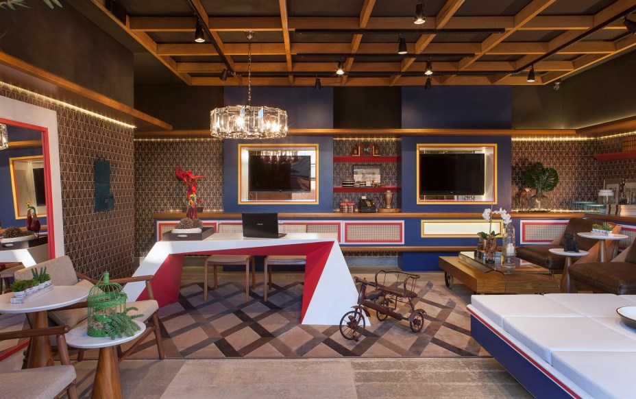 Lounge de Recepção - Lopes de Moura Studios. O espaço foi inspirado no Jockey Club de São Paulo. Os móveis trazem tons de madeira e cores primárias - vermelho, azul e amarelo. A mesa principal, desenhada exclusivamente para a mostra, é a protagonista do ambiente que abriga obras dos artistas plásticos Bia Doria e G. Comini.