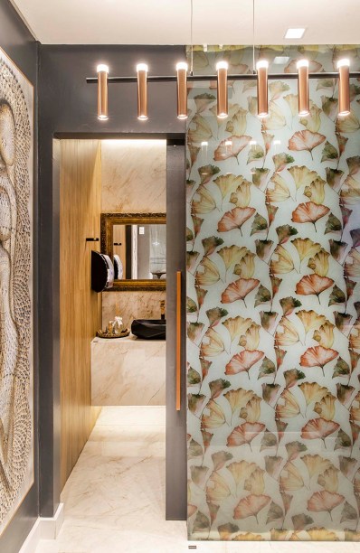 Lavabos Unissex - Janete Valladão. O espaço de 10 m² exibe iluminação moderna, uma parede com print floral protegida pelo vidro e um porcelanato que reproduz o mármore da região italiana de Sorrento no piso.