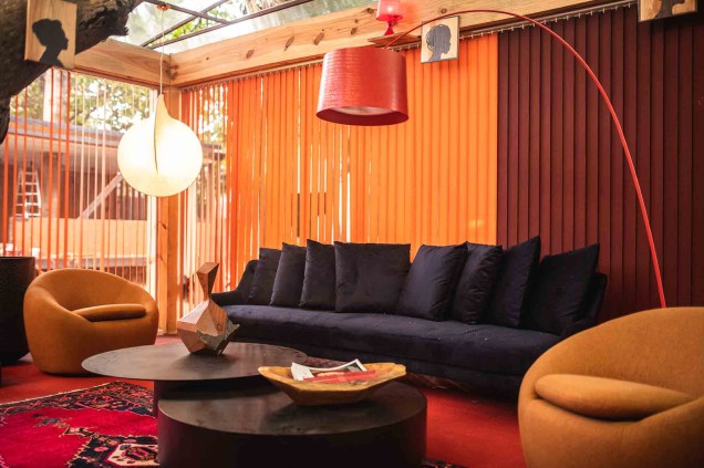 Lounge do Publisher - Roberto Pamplona Jr - CASACOR Ceará 2019. O lounge foi criado para receber convidados e visitantes, com uma locação diferente:  embaixo de frondosas árvores do local. Tons quentes entre vermelho e laranja compõem uma paleta ensolarada que aquece o local.