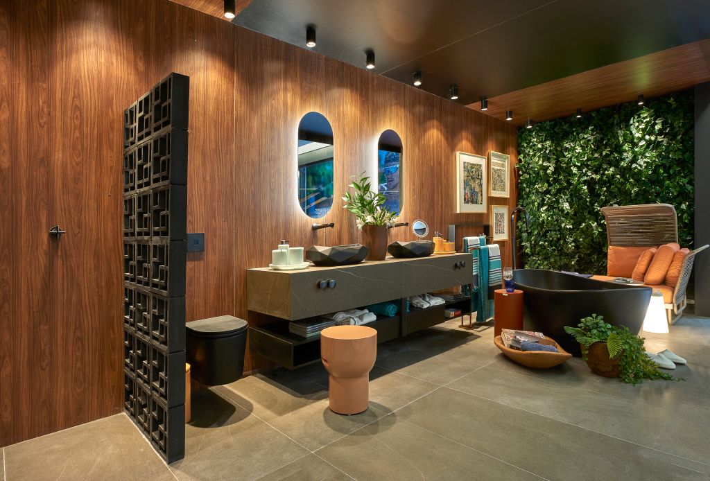 Sala de banho com chuveiro, duas pias, dois espelhos elípticos, uma banheira preta, sofá e jardim vertical