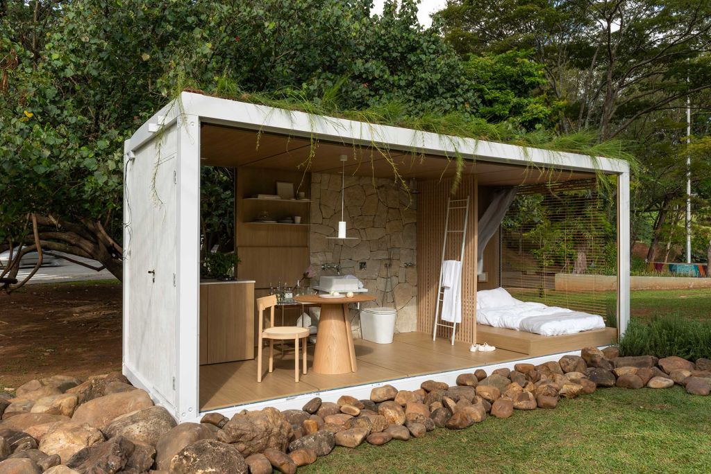 Contêiner branco, com quarto, cozinha e banheiro com mobiliário e peças em madeira. Louças em branco e o telhado verde.