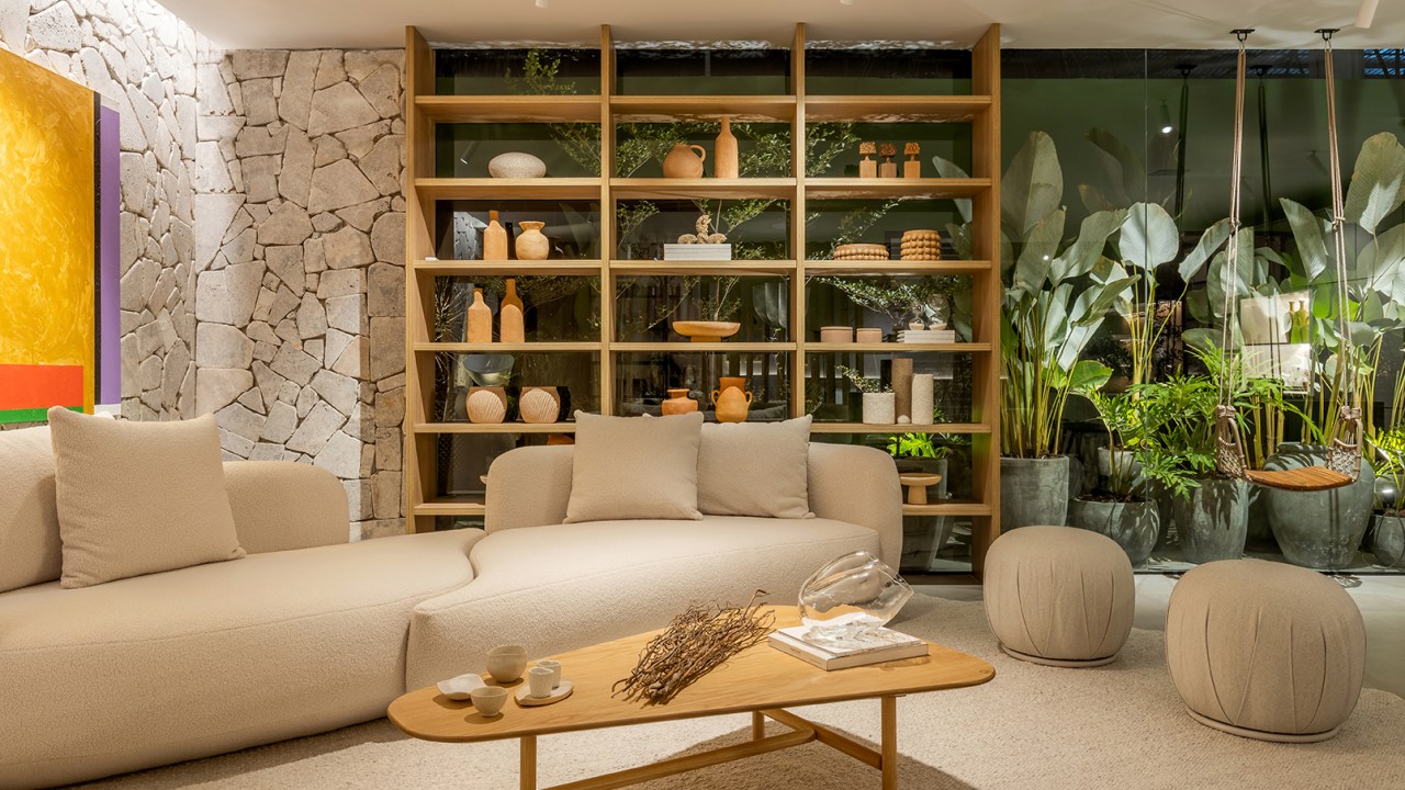 Cristiane Moussa - Refúgio Cenário. Projeto da CASACOR Goiás 2023. Na foto, sala de estar com plantas, sofá curvo e parede de pedra.