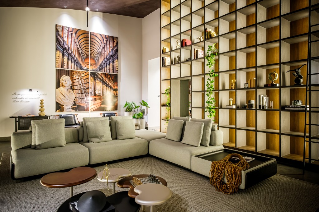 Kelly Leite - Living Principal Estar. Projeto da CASACOR Bolívia 2023. Na foto, sala de estar com estante alta e iluminada, sofá, mesa e quadros.