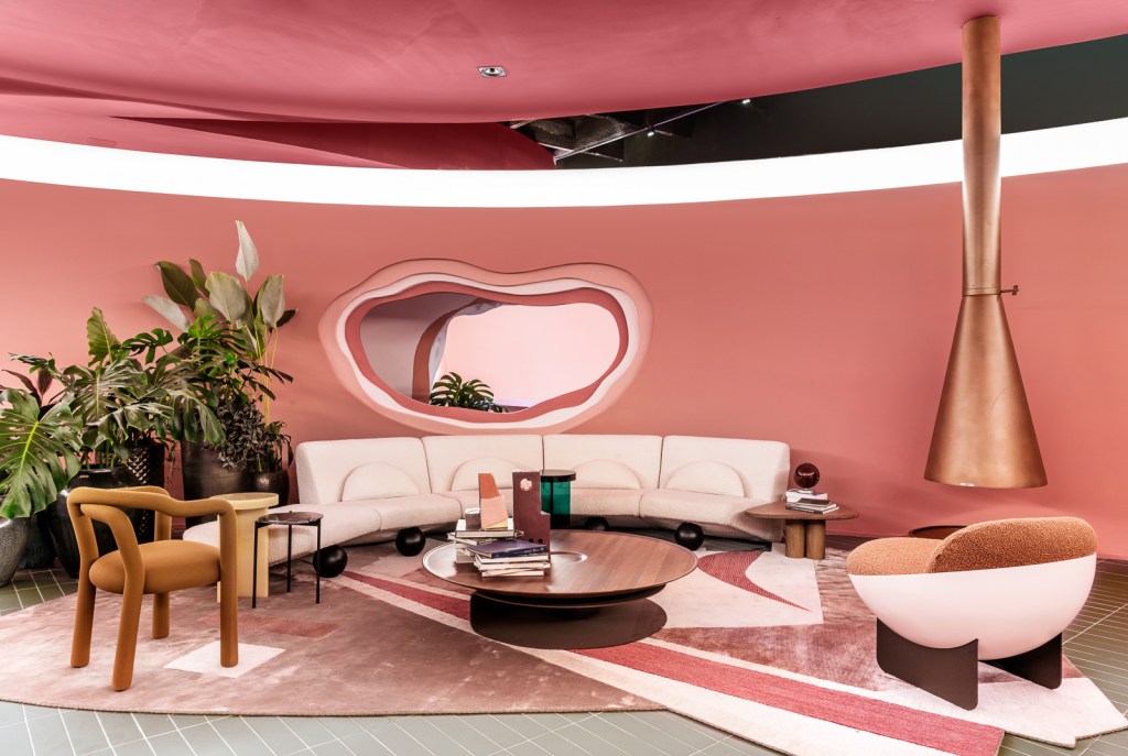 Ricardo Abreu - Casa Coral. Projeto da CASACOR São Paulo 2023. Sala com paredes rosas, sofá curvo, plantas, lareira e tapete.