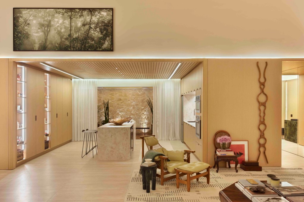 Da Hora Arquitetura - Loft Vastu. Projeto da CASACOR São Paulo 2023. Na foto, sala de estar com pé-direito duplo, estante, poltronas e tapete. Cozinha com ilha e parede de pedra.