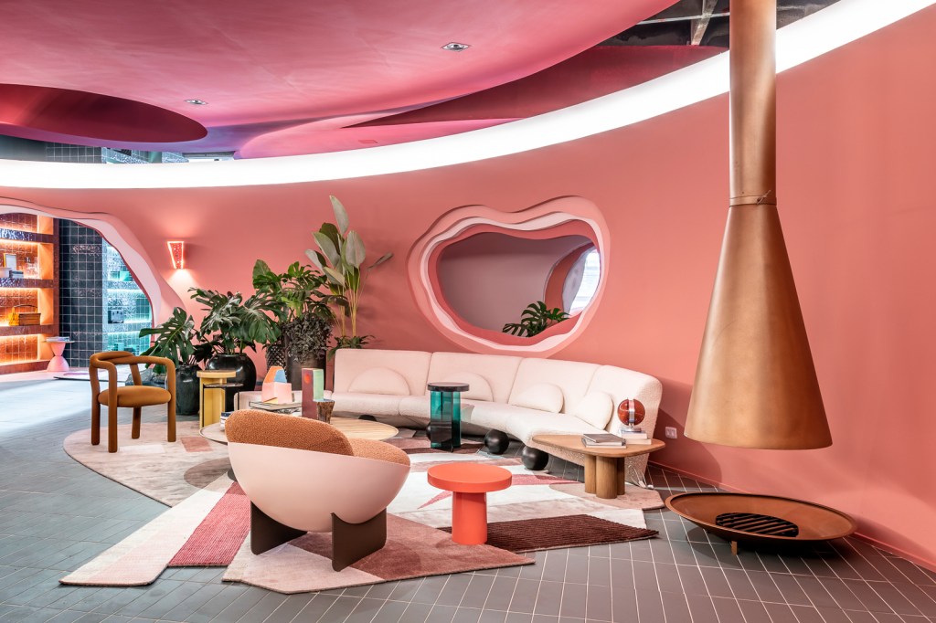 Ricardo Abreu - Casa Coral. Projeto da CASACOR São Paulo 2023. Sala com paredes rosas, sofá curvo, plantas, lareira e tapete.