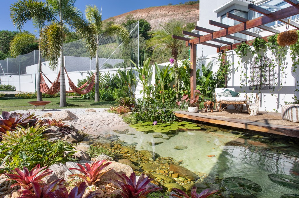 Jardim de 900 m² tem lago com peixes, praia de areia branca e pomar. Projeto de Ana Veras e Bernardo Vieira. Na foto, jardim com lago, pergolado, prainha de areia e rede.