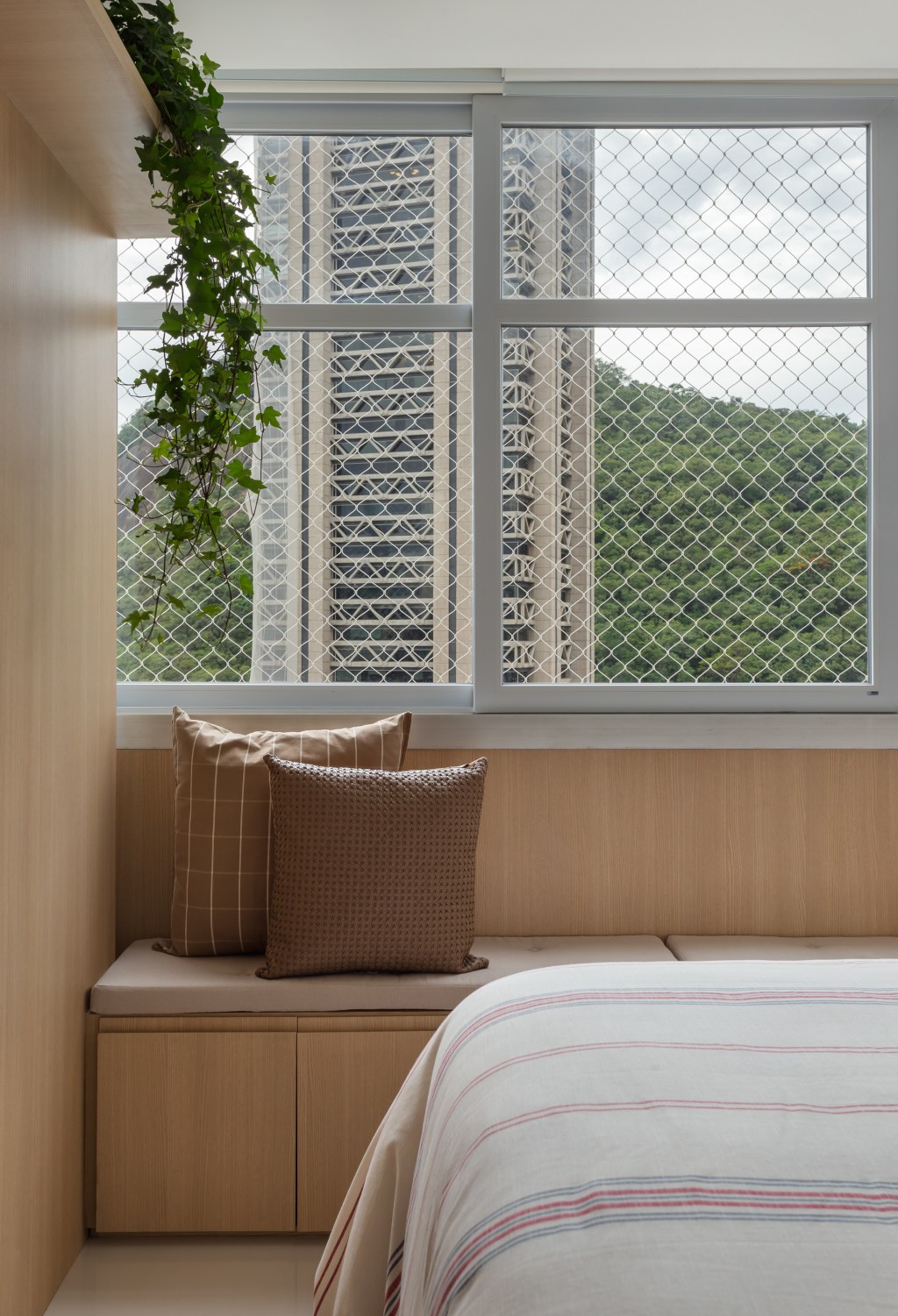 Estante multiuso setoriza a área social deste apê de 90 m². Na foto, quarto de casal com banco na janela e vista para natureza.