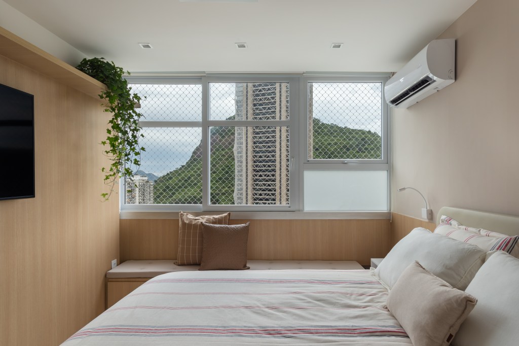 Estante multiuso setoriza a área social deste apê de 90 m². Na foto, quarto de casal com vista para a natureza, parede de madeira e banco na janela.