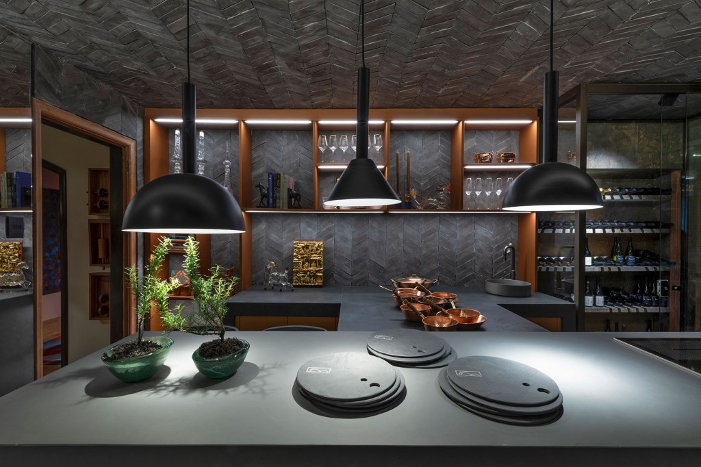 Andréa Buratto - Cozinha cave. Projeto da CASACOR Minas Gerais 2023. Na foto, cozinha com bancada, de refeições, mesa, luminárias e estante e adega.