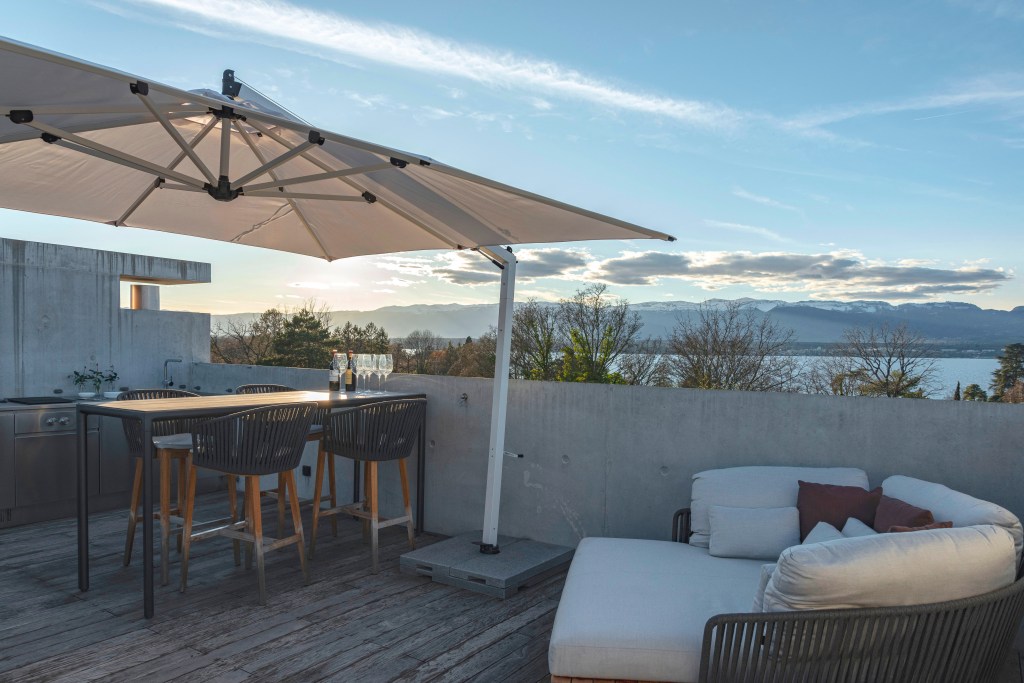 David Bastos projeta apartamento na Suíça repleto de design assinado. Na foto, varanda gourmet com ombrelone, sofá e mesa.