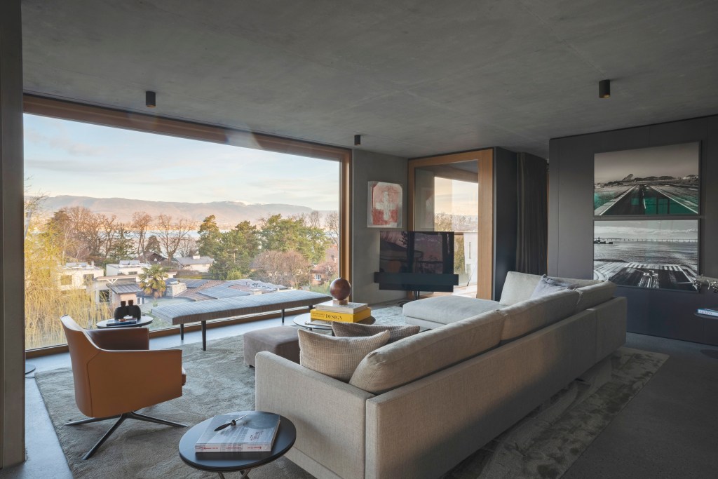 David Bastos projeta apartamento na Suíça repleto de design assinado. Na foto, sala de estar com parede de vidro, sofa cinza e tapete.