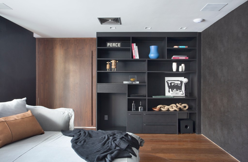 Décor masculino e repleto de design assinado marcam este apê de 350 m². Projeto Gabriela Eloy. Na foto, sala de TV com estante preta, cama, piso de madeira e parede preta.