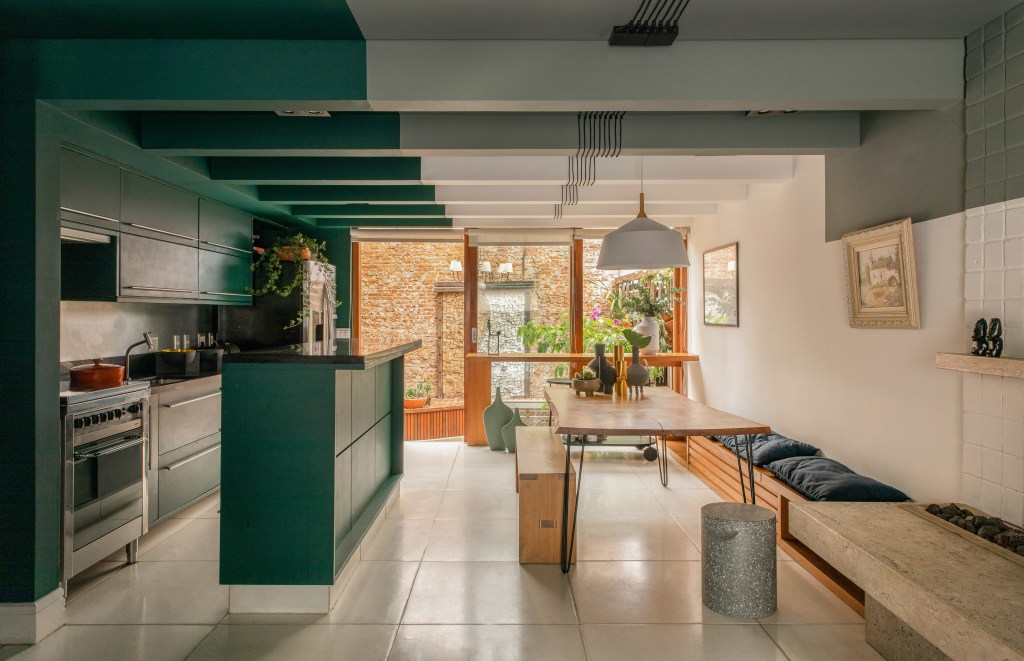 Degradê de tons de verde setoriza esta casa de vila em São Paulo. Projeto de Ricardo Abreu. Na foto, sala e cozinha integradas com paredes e marcenaria verde, banco na sala de jantar e vista para o jardim.