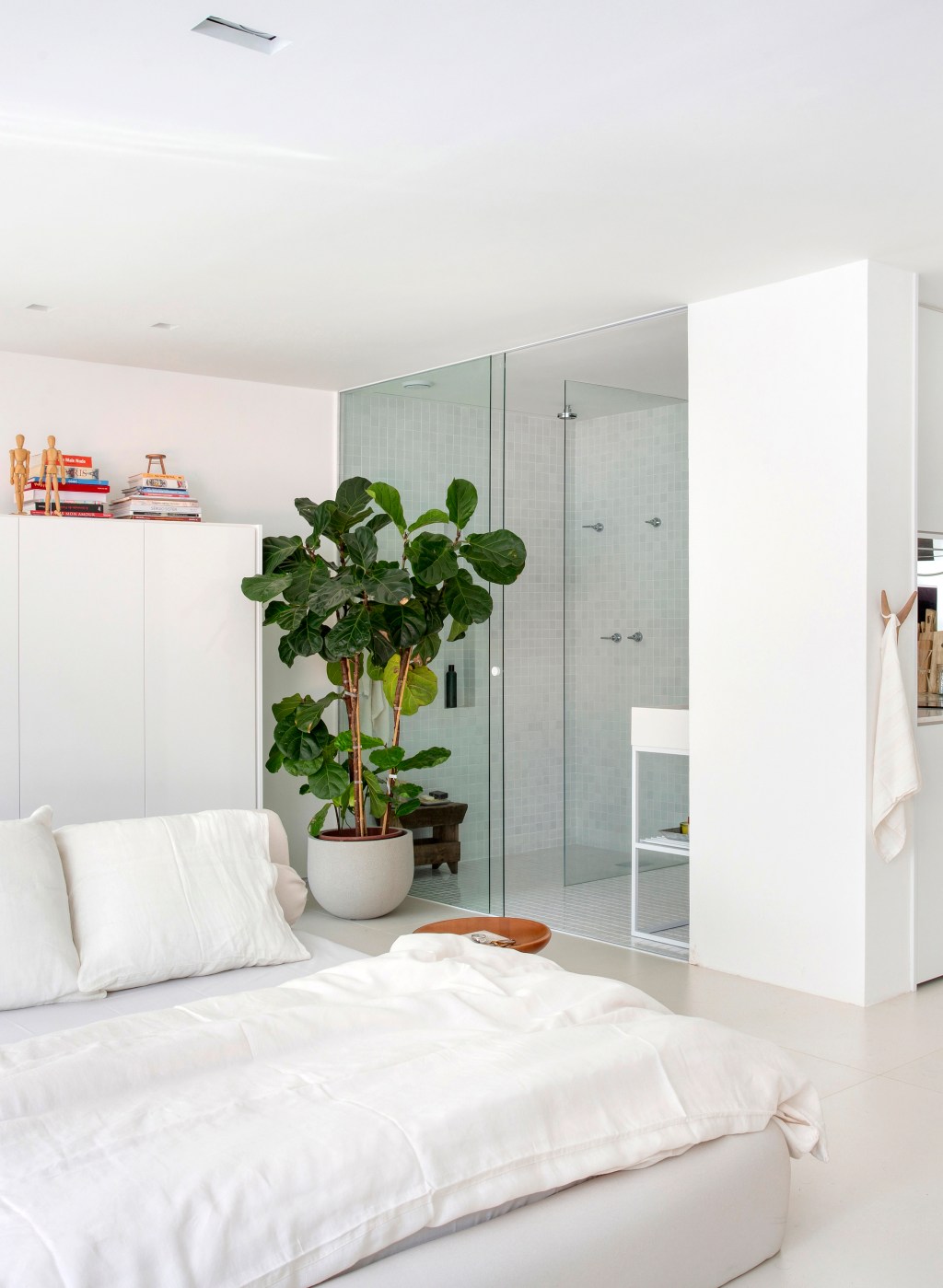 Loft de 40 m² ganha projeto minimalista que une branco e madeira. Projeto de Diego Raposo e Manuela Simas. Na foto quarto com banheiro integrado. Vaso de plantas e cama.