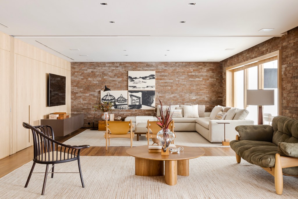 Parede de tijolos e painéis de madeira trazem acolhimento ao apê de 310 m². Projeto Très Arquitetura. Na foto, sala de estar com parede de tijolinhos e madeira e varanda integrada.