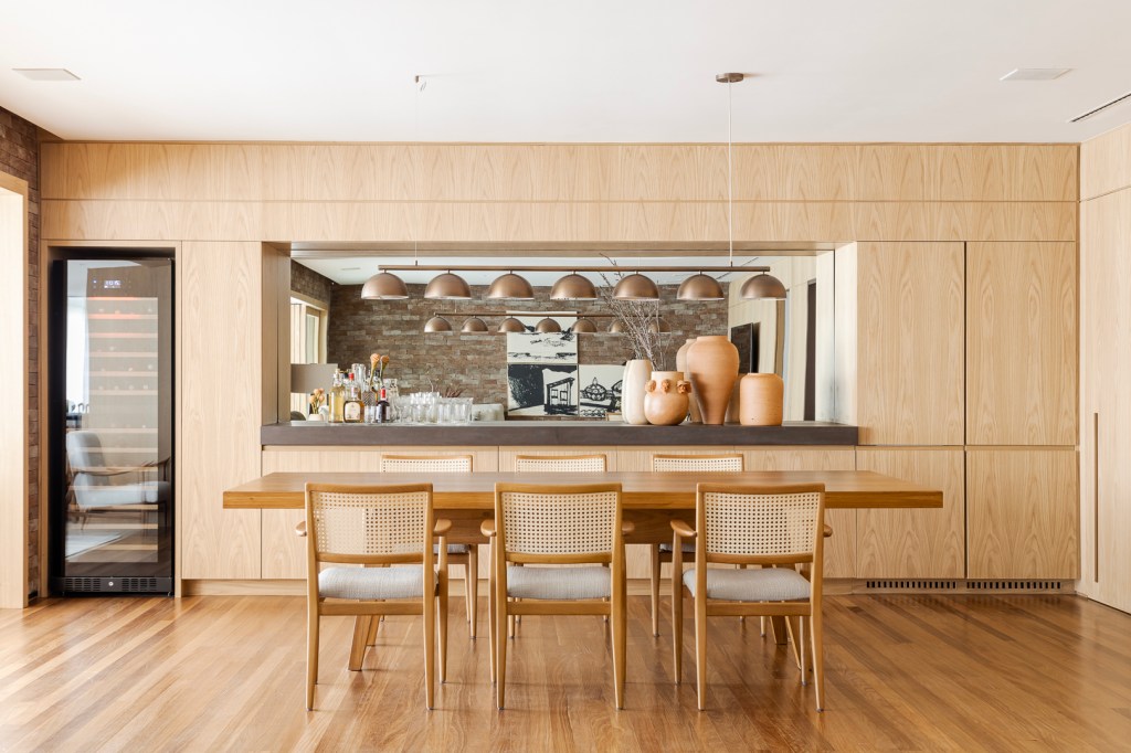 Parede de tijolos e painéis de madeira trazem acolhimento ao apê de 310 m². Projeto Très Arquitetura. Na foto, sala de estar com parede de tijolinhos e madeira e sala de jantar integrada.