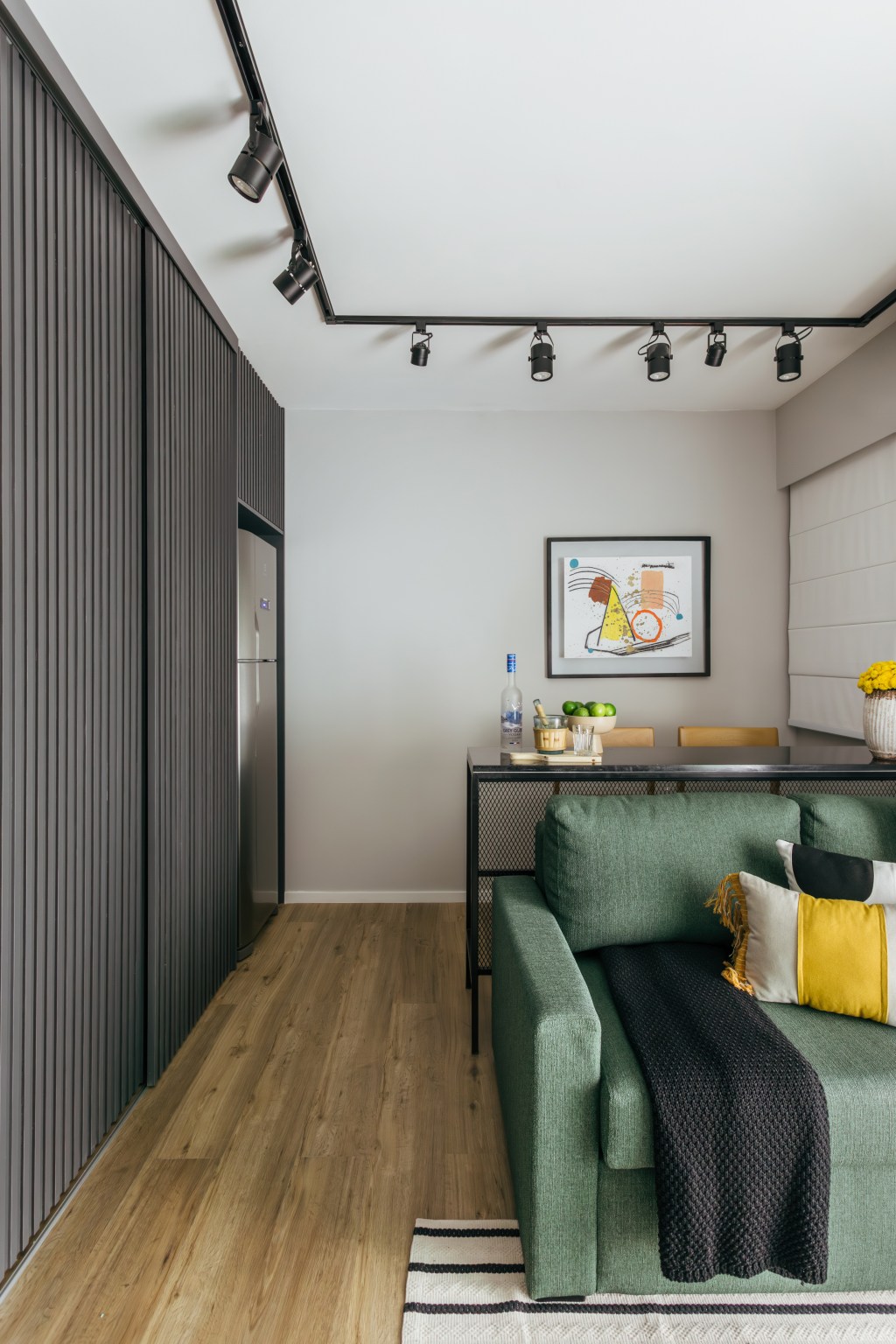 Cozinha se esconde dentro do armário neste apê de 35 m², Projeto de Ikeda Arquitetura. Na foto, sala de estar com bancada de serralheria, cozinha camuflada e sofá verde.