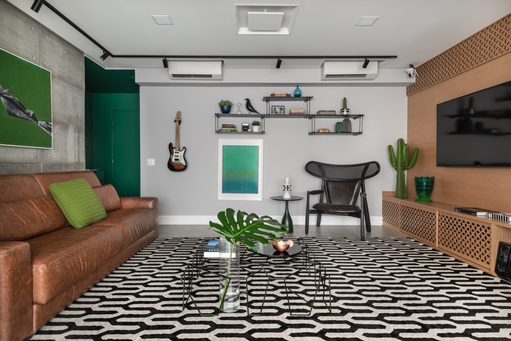 Painel retrátil esconde a churrasqueira neste apê descolado de 120 m². Projeto de Beatriz Quinelato. Na foto, sala de estar com parede verde, sofá de couro e tapete estampado.
