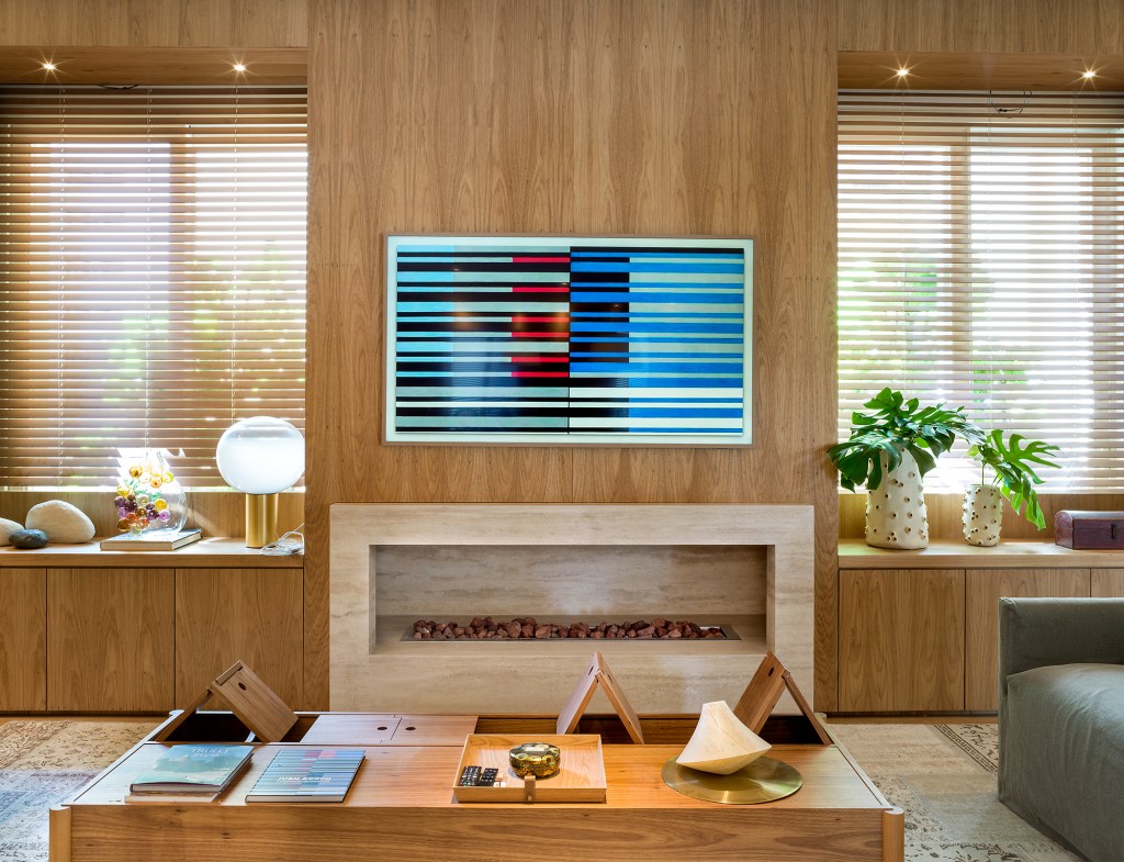 Paola Ribeiro assina apartamento de 810 m² repleto de arte. Na foto, sala de estar com quadro, lareira e parede de madeira.