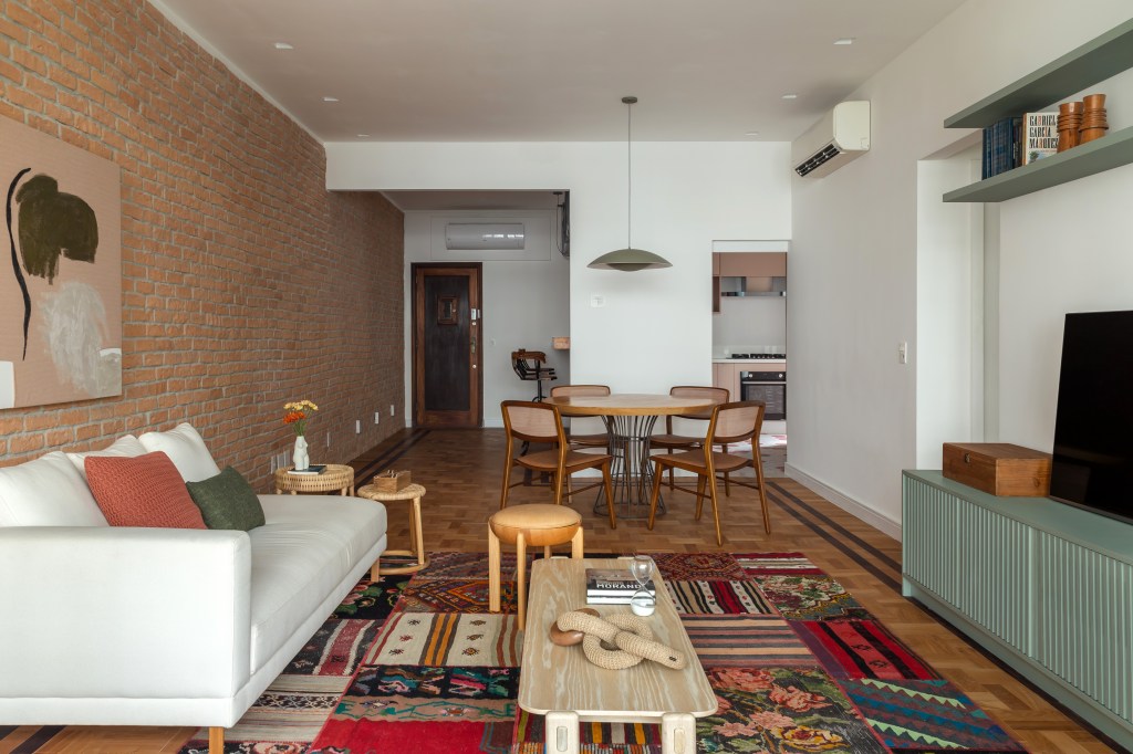 Apê de 145 m² ganha charme com tijolinhos, ladrilho hidráulico e patchwork. Na foto, sala com parede de tijolinhos, tapete de patchwork e aparador. Sala de jantar com mesa redonda.