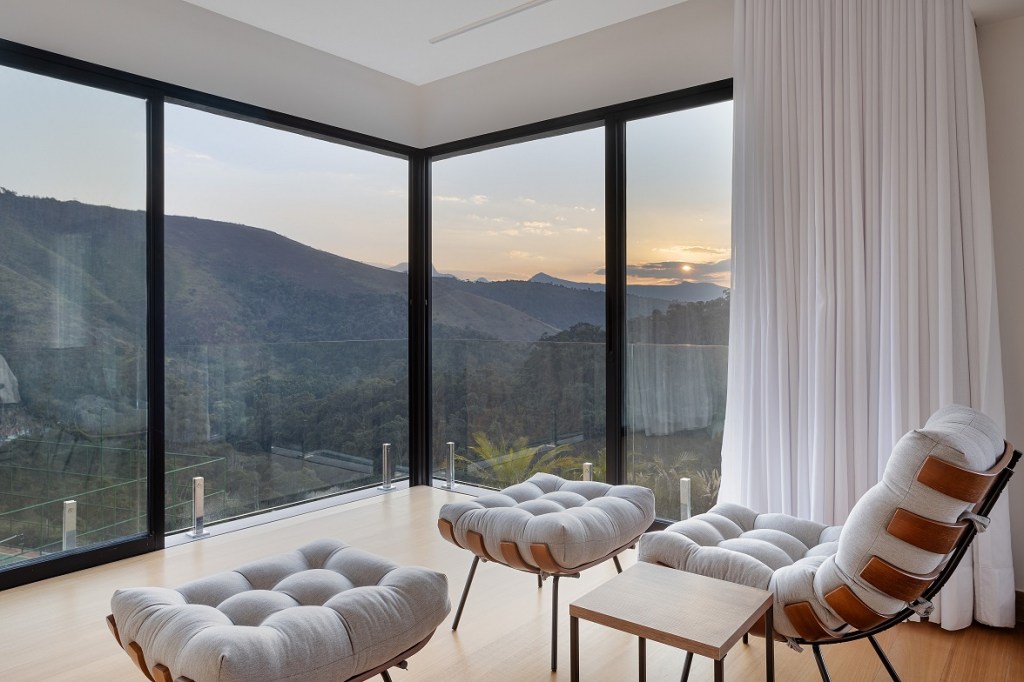 Casa de 1200 m² na serra tem clima de clube de férias. Projeto de Rafael Mirza. Na foto, Na foto, quarto com parede de vidro e vista para a montanha.