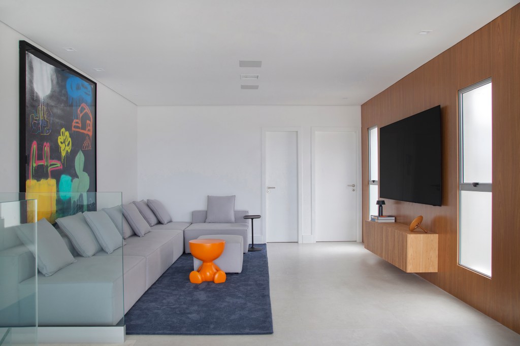 Cobertura de 400 m² feita para gêmeos é repleta de design e tecnologia. Projeto de Tom Castro. Na foto, sala de TV com parede de madeira e sofá cinza.