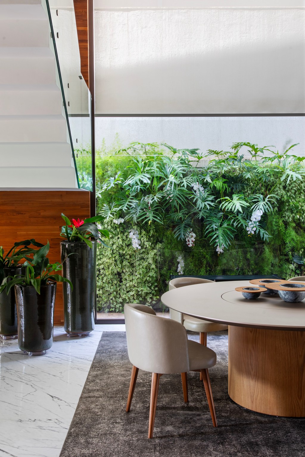 Jardim tropical traz mix de folhagens, texturas e vegetação. Projeto de Horto Girassol. Na foto, sala de jantar com parede verde, mesa redonda e cadeiras.