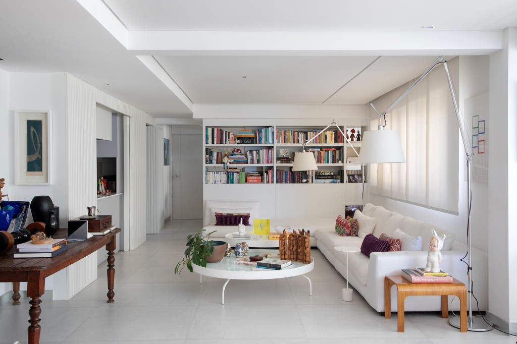 Conheça o apê da arquiteta Ana Moura, do elenco da CASACOR Pernambuco. Na foto, sala de estar com estante branca, cozinha separada com porta de correr e sofá.