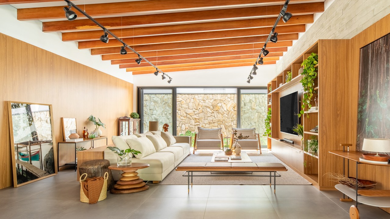 Pensada para veraneio, casa em meio ao verde vira o lar oficial da família. Projeto de Pietro Terlizzi. Na foto, sala de tv com estante de madeira e ripas no teto. Muro de pedra.