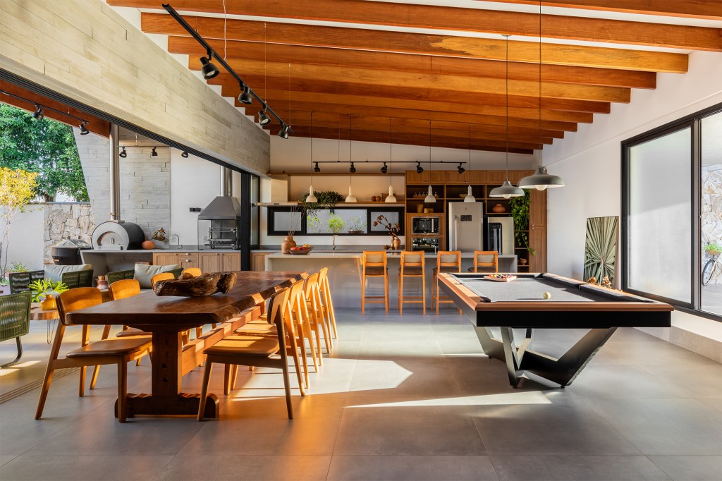 Pensada para veraneio, casa em meio ao verde vira o lar oficial da família. Projeto de Pietro Terlizzi. Na foto, varanda e sala integradas com mesa de jantar, mesa de sinuca e cozinha.