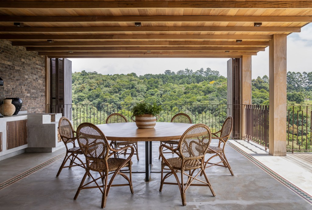 Casa sustentável e integrada à natureza fica em frente ao campo de golfe. Projeto de In House Arquitetura. Na foto, varanda com mesa de jantar e forno a lenha.