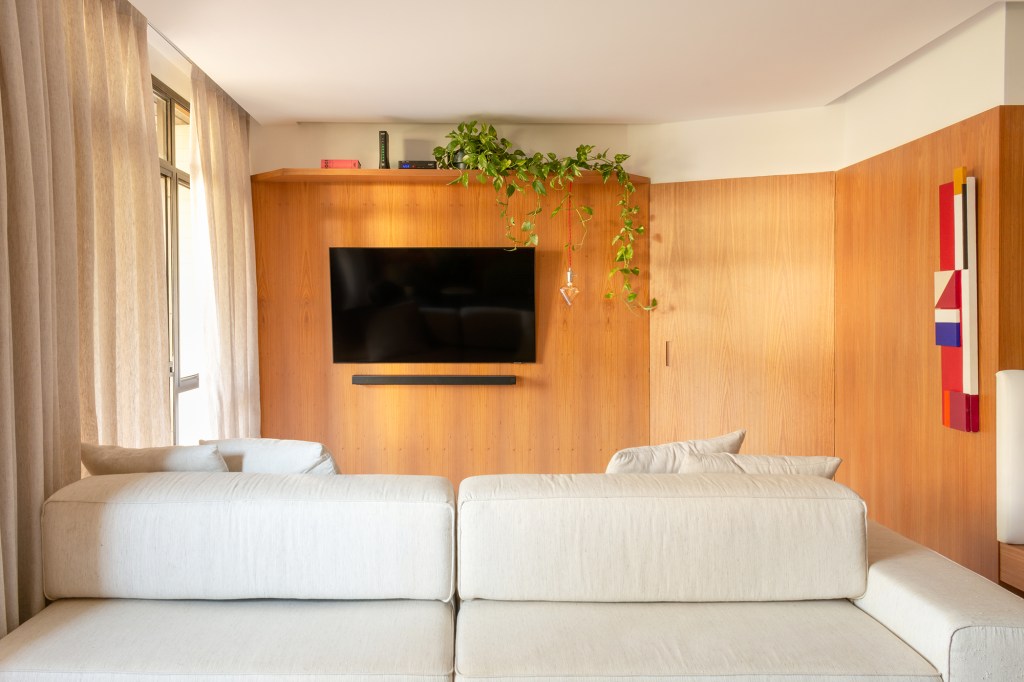 A madeira é a protagonista deste apartamento de 117 m² na Gávea. Projeto de Mariana Monnerat, Na foto, sala com tv, painel de madeira e sofá ilha.