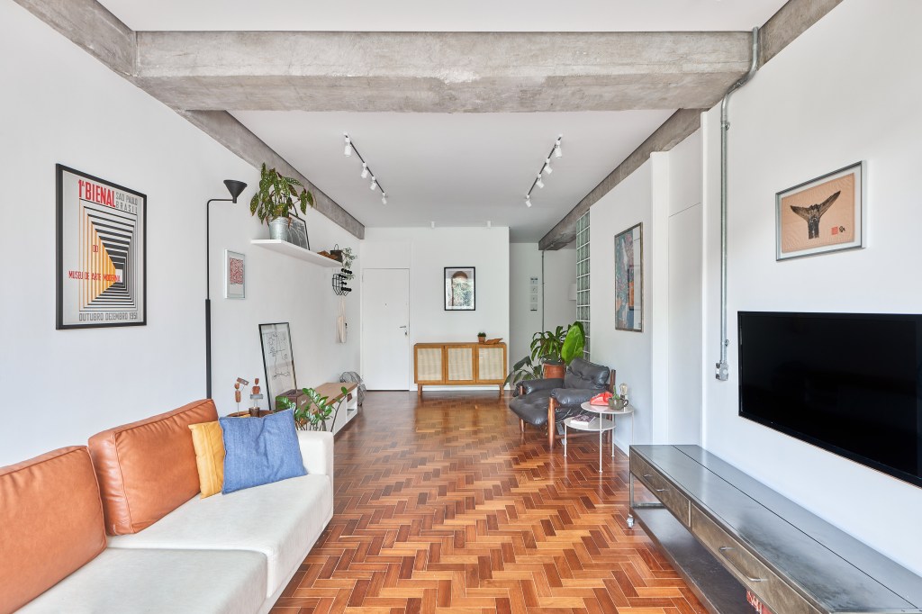 Apartamento de 98 m² ganha décor contemporâneo, mas com toques de brutalismo. Projeto de Hugo Rapizo. Na foto, sala com sofá, quadro, piso de taco e varanda com rede.