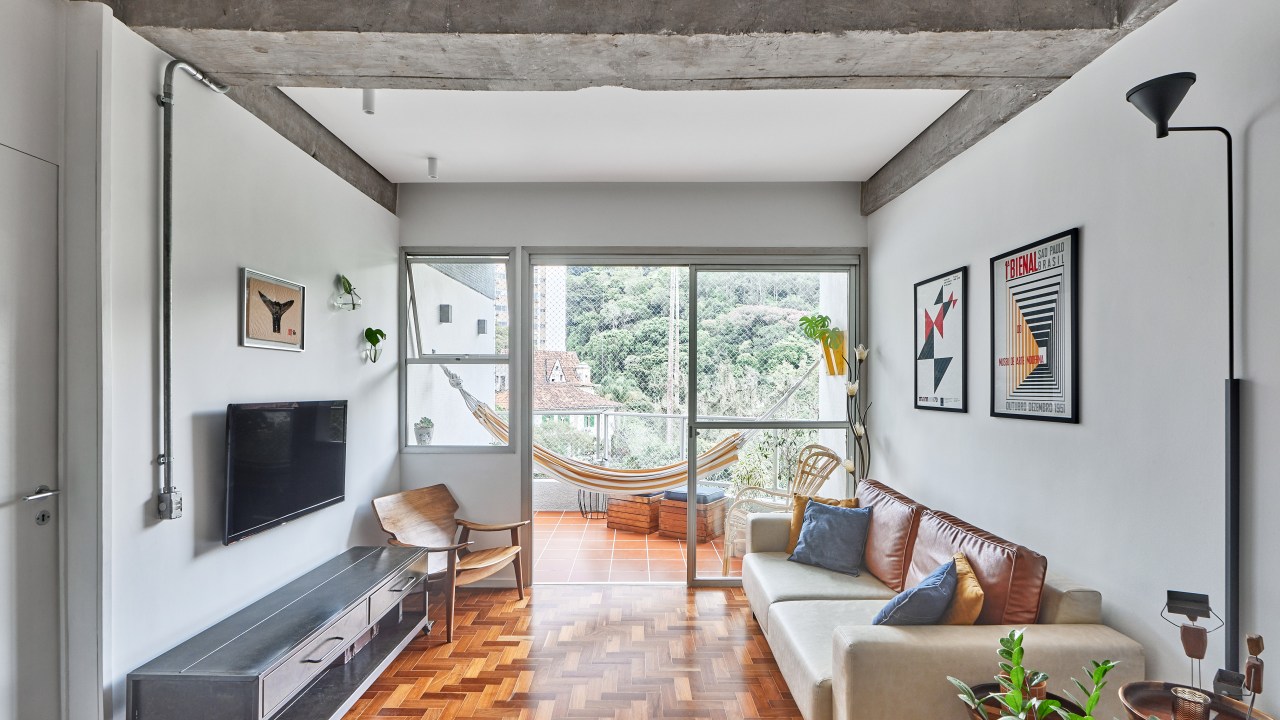 Apartamento de 98 m² ganha décor contemporâneo, mas com toques de brutalismo. Projeto de Hugo Rapizo. Na foto, sala com sofá, quadro, piso de taco e varanda com rede.