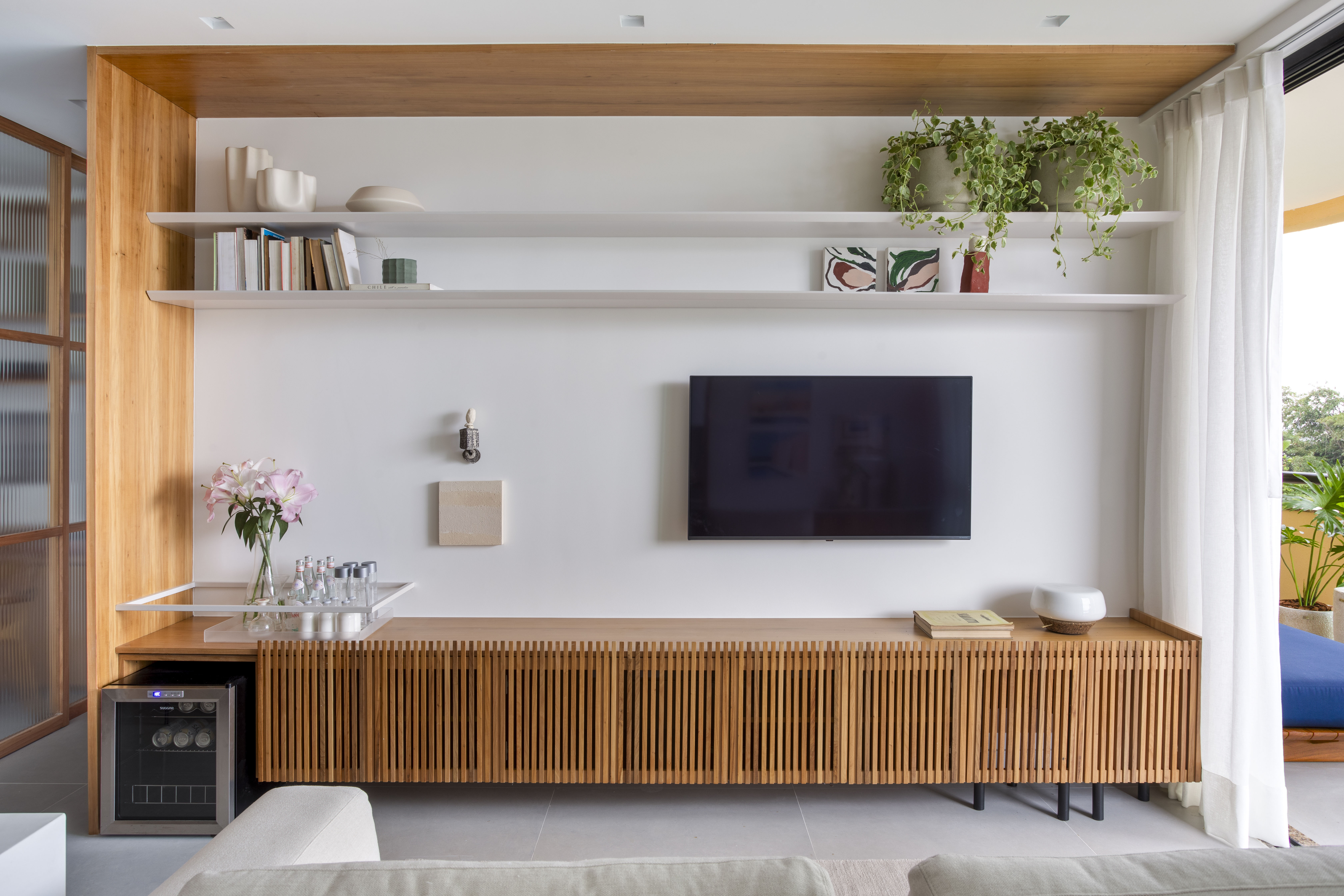 Branco e madeira criam décor cozy em apartamento de 70 m². Projeto de Rafael Ramos. Na foto, sala de tv com aparador ripado e prateleiras.